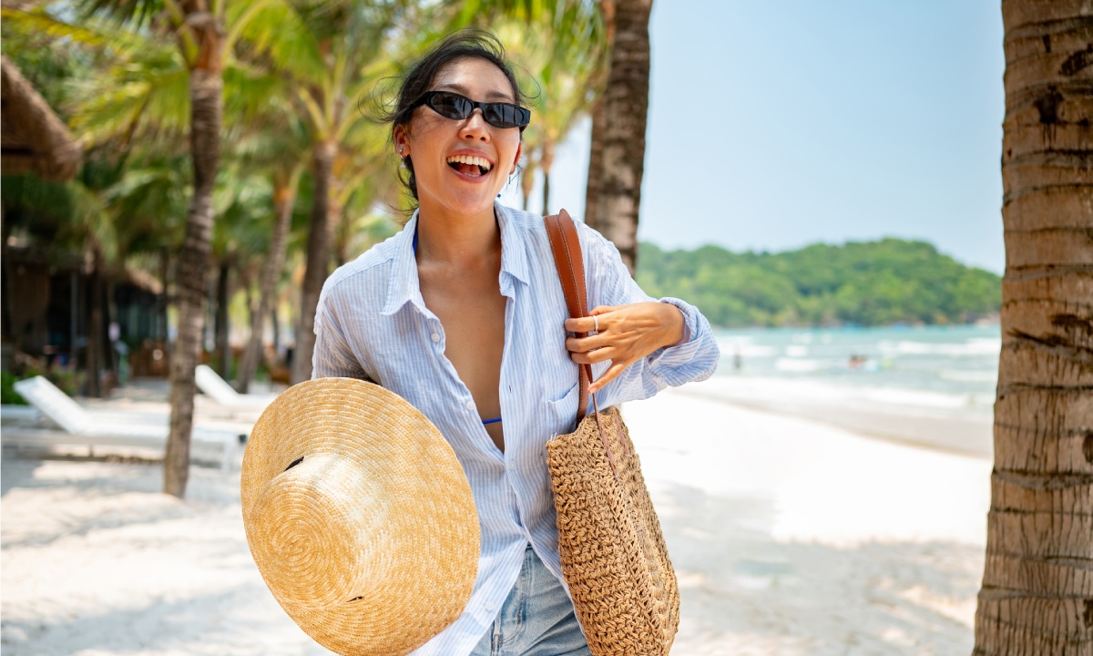 A woman with sunglasses, hat and beach bag on a palm-lined beach
