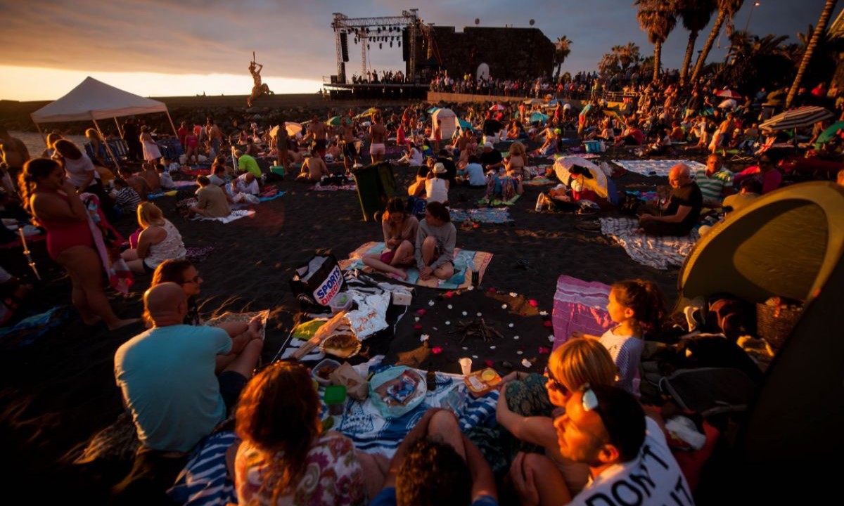 San Juan Night at Playa Jardín, Puerto de la Cruz, Tenerife, with people gathered on the beach and a stage at sunset