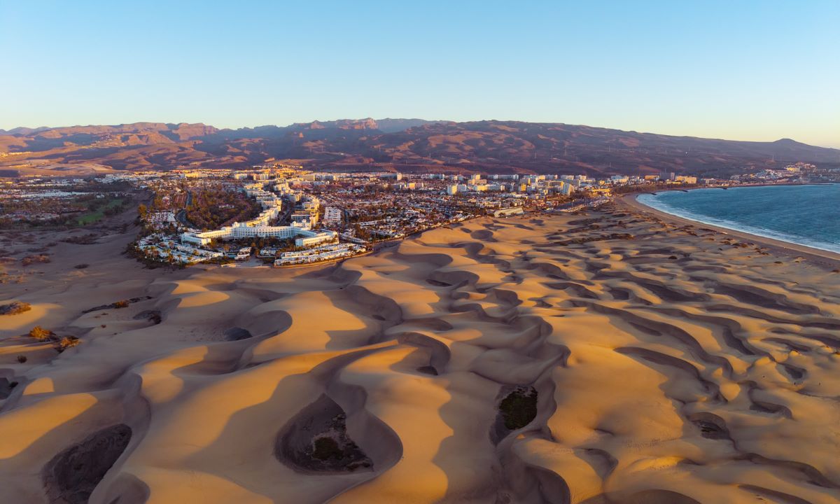 Vista aérea de las espectaculares Dunas de Maspalomas en Gran Canaria al atardecer, con hoteles y montañas al fondo.