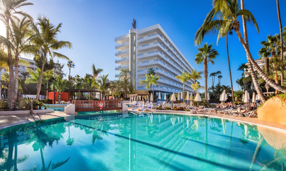 Piscina principal con aguas cristalinas del hotel Gran Canaria Princess rodeada de palmeras y hamacas bajo un cielo despejado.