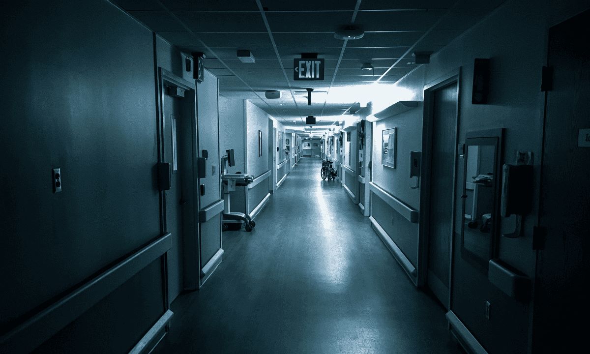 A dark, gloomy hospital corridor with cold, bluish lighting and a wheelchair at the far end.