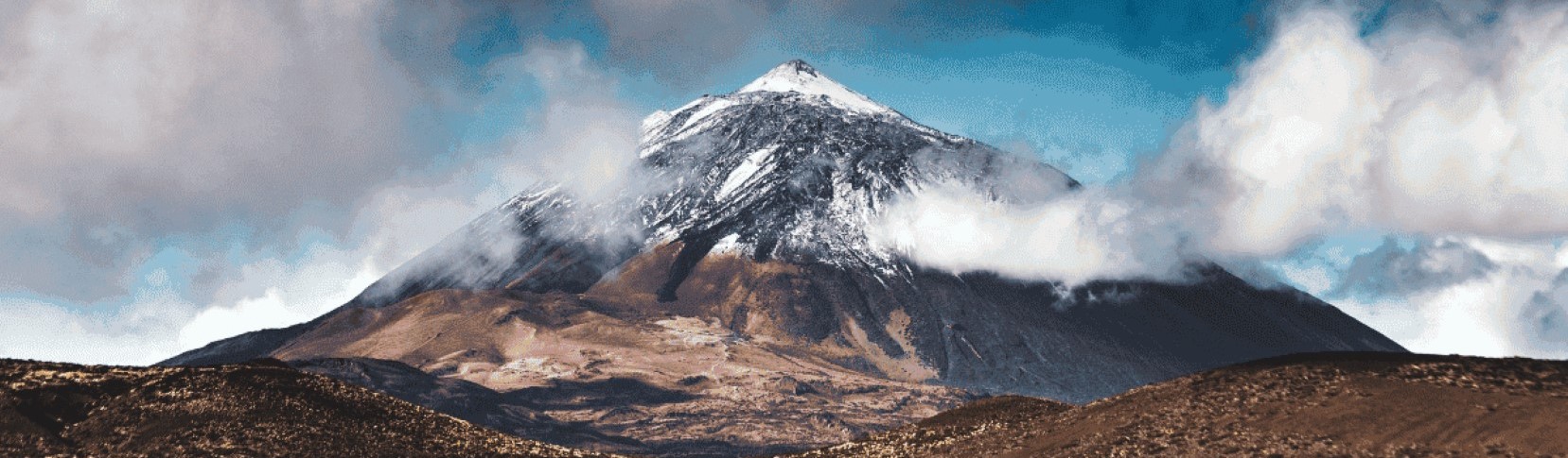 The snow-capped peak of Mount Teide surrounded by dark clouds beneath a blue sky, said to be Guayota's home in Tenerife