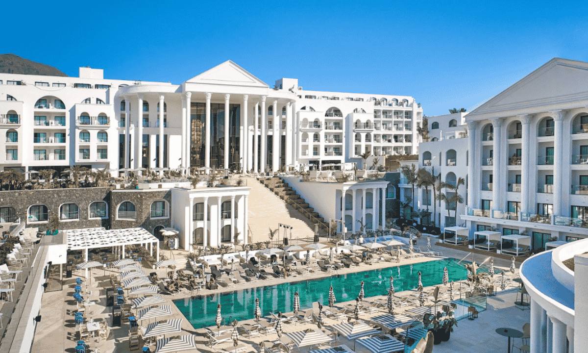 Front view of the outdoor pool surrounded by sun loungers and parasols, facing the striking white classic-style façade of Hotel Princess Inspire Tenerife in Costa Adeje.