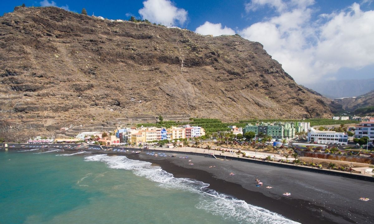 Panorámica aérea de la Playa de Tazacorte con sombrillas en su arena negra, un mar calmado de tonos turquesas y las coloridas edificaciones del pueblo frente a un gran acantilado.