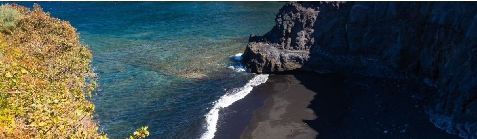 Vista desde lo alto de la Playa de La Zamora en Fuencaliente, mostrando una cala de arena negra rodeada de imponentes acantilados oscuros y formaciones rocosas en el mar azul.