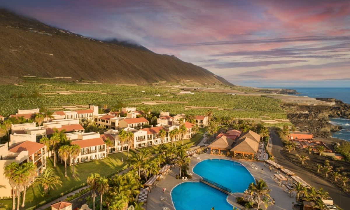 Vista aérea de Esencia de La Palma en el hotel La Palma Princess con piscinas azules, plantaciones de plátanos y acantilados al atardecer.