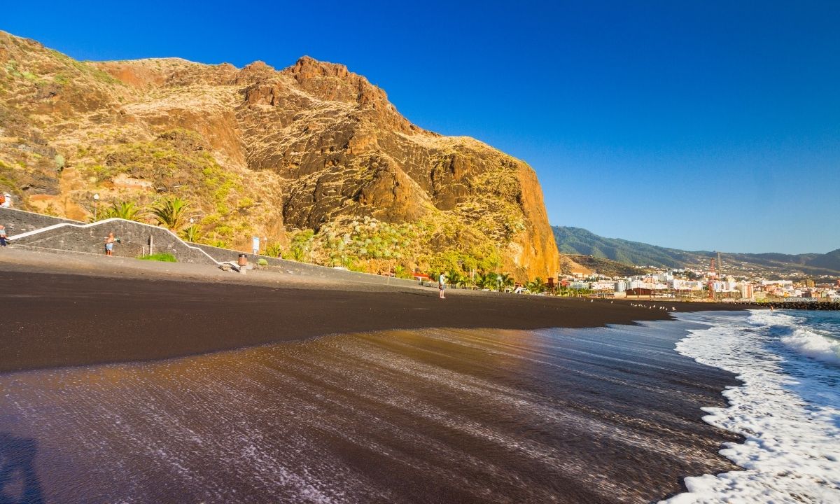Orilla de la Playa de Bajamar mostrando el contraste de la espuma blanca del mar sobre la extensa arena negra volcánica, flanqueada por un enorme acantilado rocoso bajo un cielo despejado.