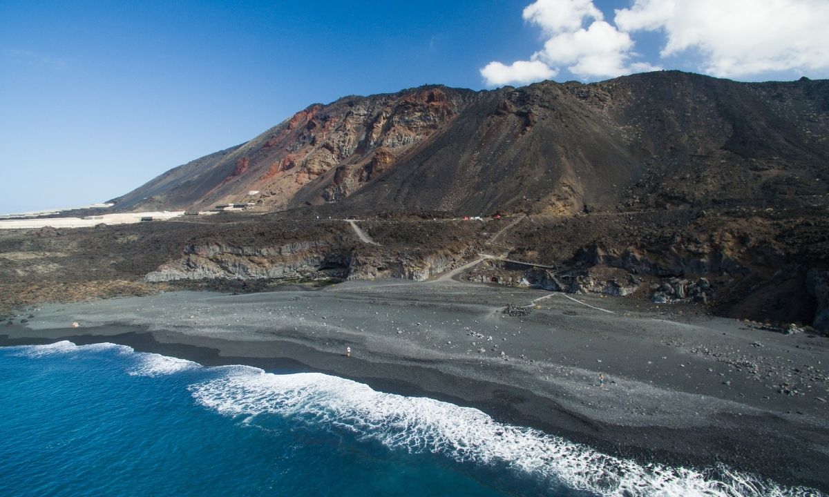 Alt Text: Vista aérea de la Playa de Echentive con su orilla de arena negra y picón, rodeada por un extenso y árido paisaje volcánico de coladas de lava bajo un cielo azul.