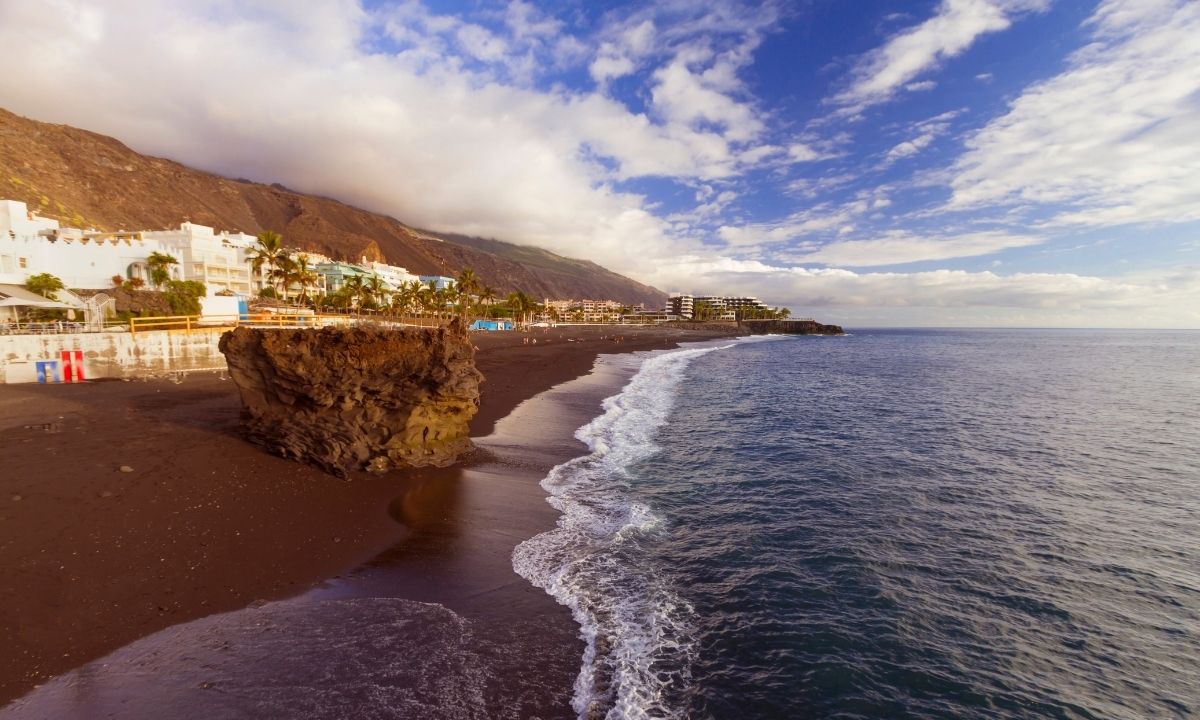 Playa de Puerto Naos con su característica arena negra volcánica, rocas en la orilla, edificios costeros y palmeras bajo un cielo parcialmente nublado en La Palma.