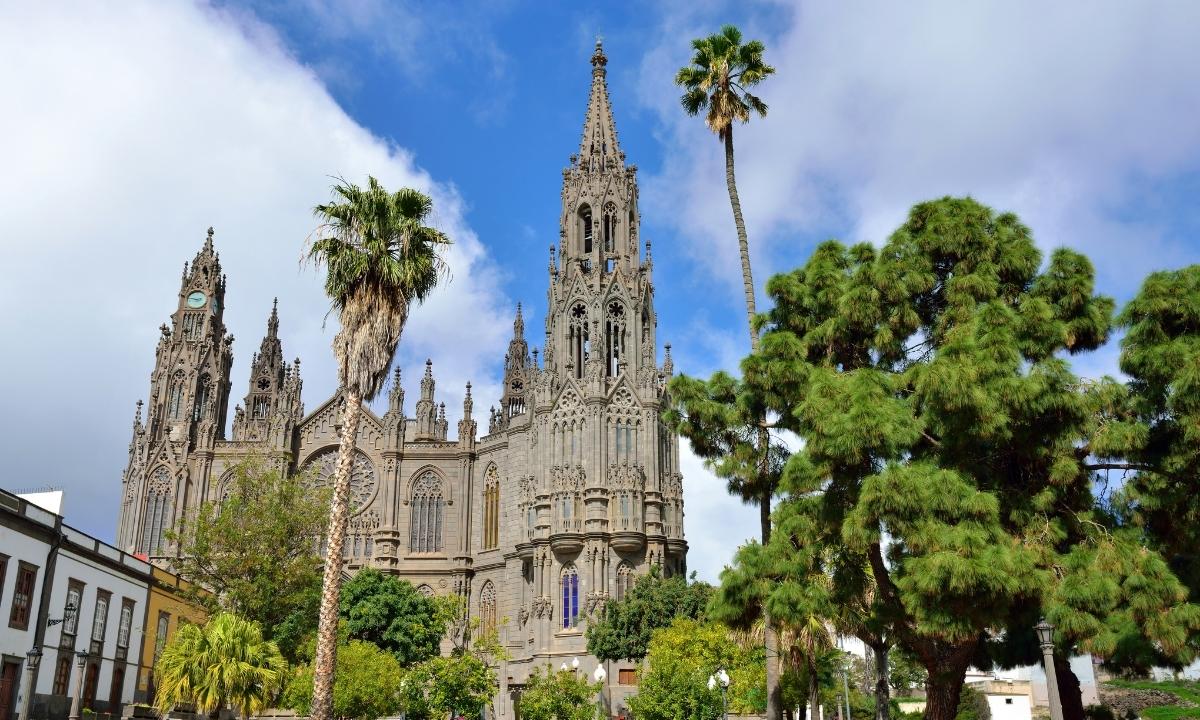 Basílica de Arucas, Gran Canaria, con palmeras y arquitectura neogótica bajo el cielo azul.