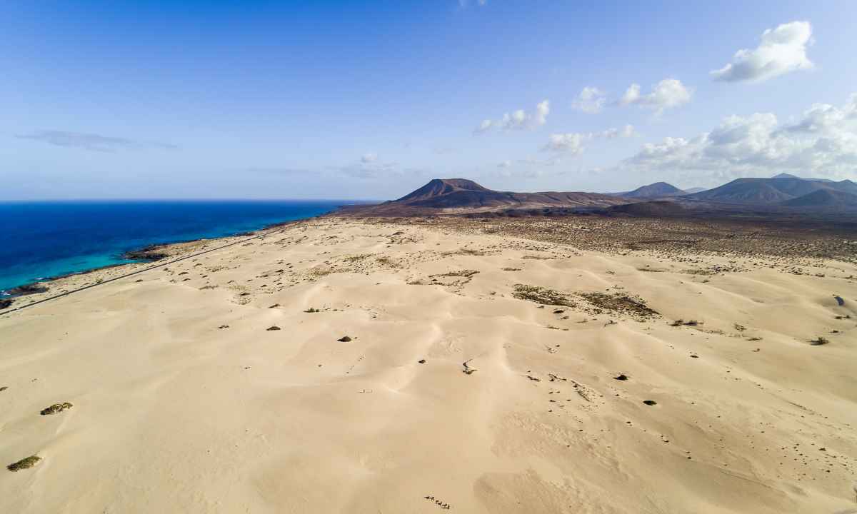 Vista aérea de las dunas de arena dorada en Corralejo junto a la costa y montañas al fondo.